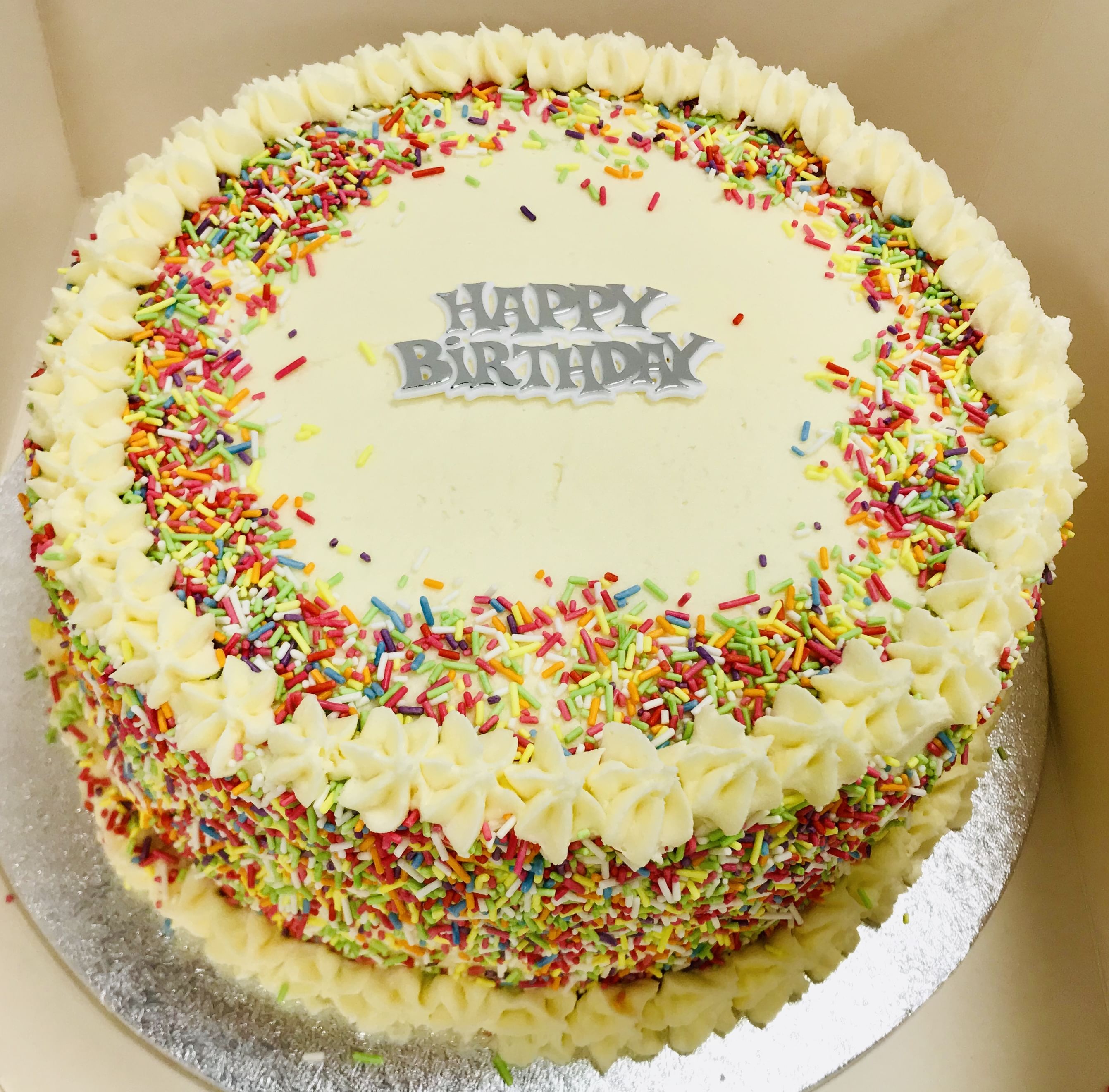 A round, frosted celebration cake adorned with multicolored sprinkles on the top and sides. The Sprinkles Cake has a white buttercream border, and 'Happy Birthday' is written in gray icing at the center on a white frosting background. It sits on a silver cake board.